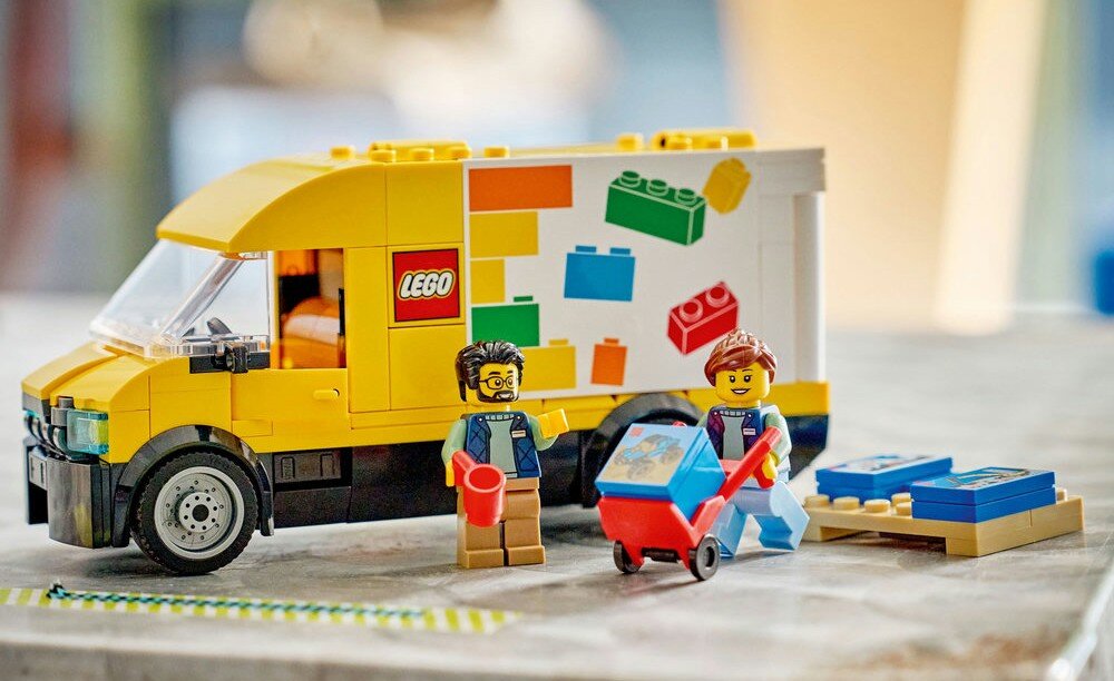 Zestaw LEGO przedstawia żółtą furgonetkę z logo LEGO oraz dwie figurki postaci z akcesoriami, takimi jak taczka i pudełka z klockami.