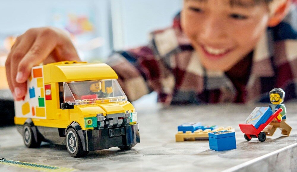 Na obrazie widoczna jest żółta furgonetka z klocków LEGO oraz figurka z wózkiem i klockami. W tle znajduje się osoba w koszuli w kratę, która bawi się furgonetką.