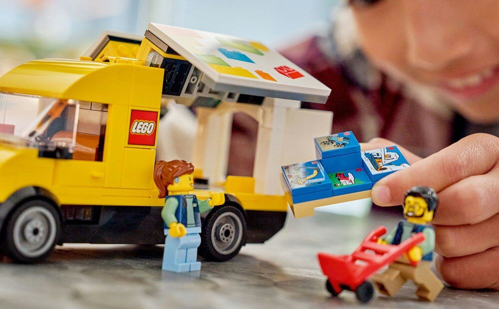 Na obrazie widoczny jest żółty furgon LEGO z otwartym dachem, obok którego znajdują się dwie figurki LEGO. Na furgonie widnieje logo LEGO.
