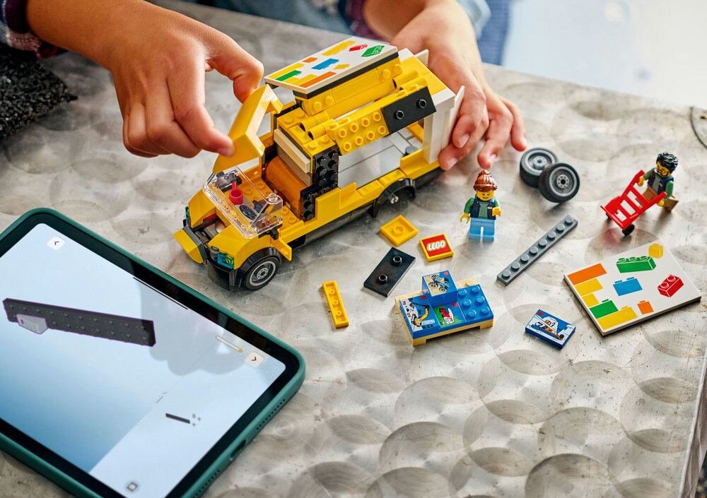 Na stole znajduje się żółta furgonetka z klocków LEGO oraz kilka rozłożonych elementów i figurek. Obok leży tablet z wyświetlonym obrazem instrukcji budowy LEGO.