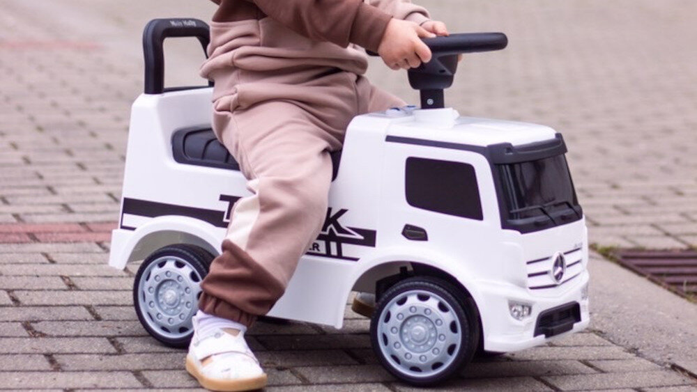 Dziecko siedzi na białym jeździku przypominającym ciężarówkę z logo w kształcie gwiazdy na przodzie. Na boku pojazdu widoczny jest napis \'TRUCK\'.