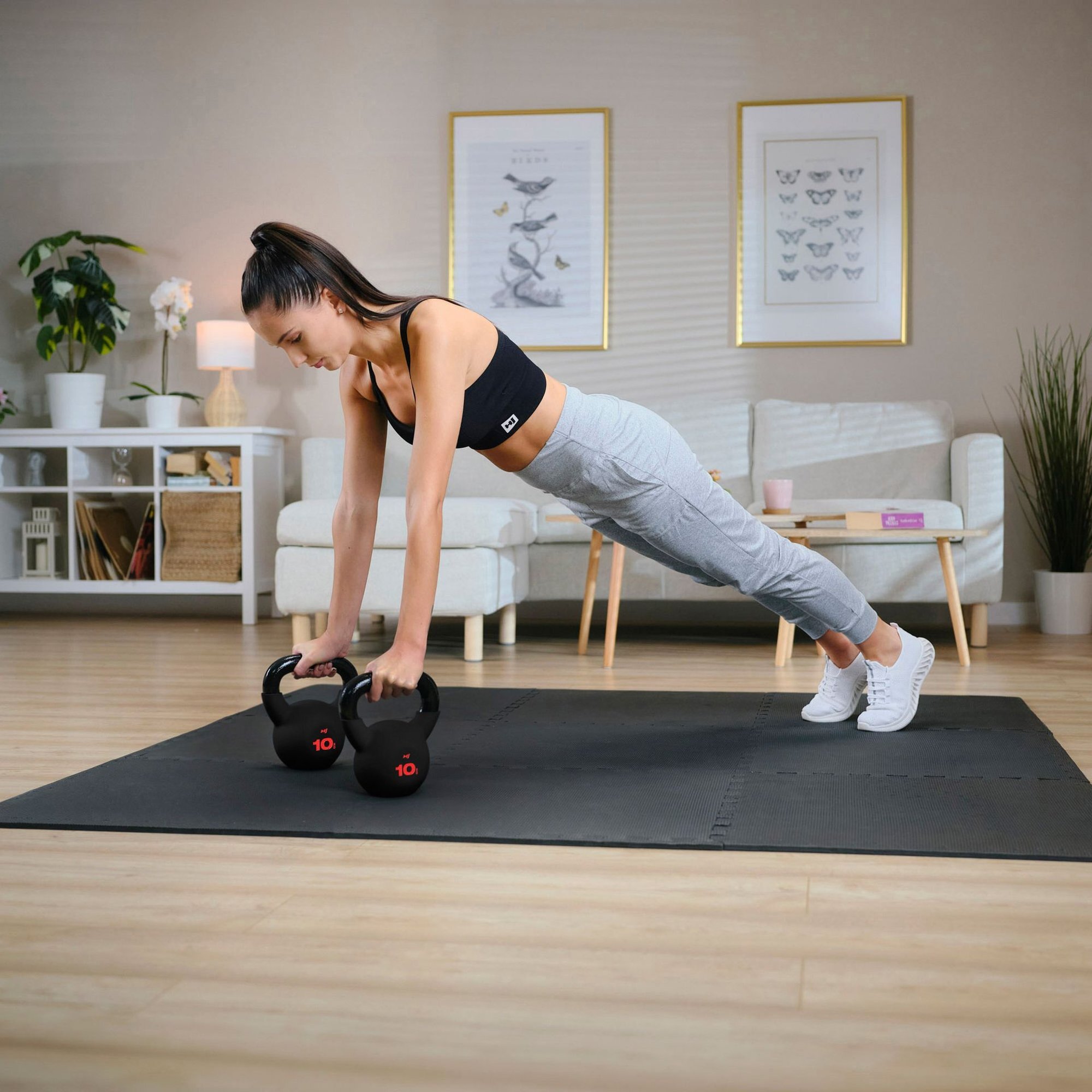 Osoba wykonuje ćwiczenie w pozycji deski, opierając się na dwóch żeliwnych odważnikach kettlebell o wadze 10 kg z widocznym napisem \'10\'. W tle znajduje się jasne wnętrze z roślinami, sofą i obrazami na ścianie.