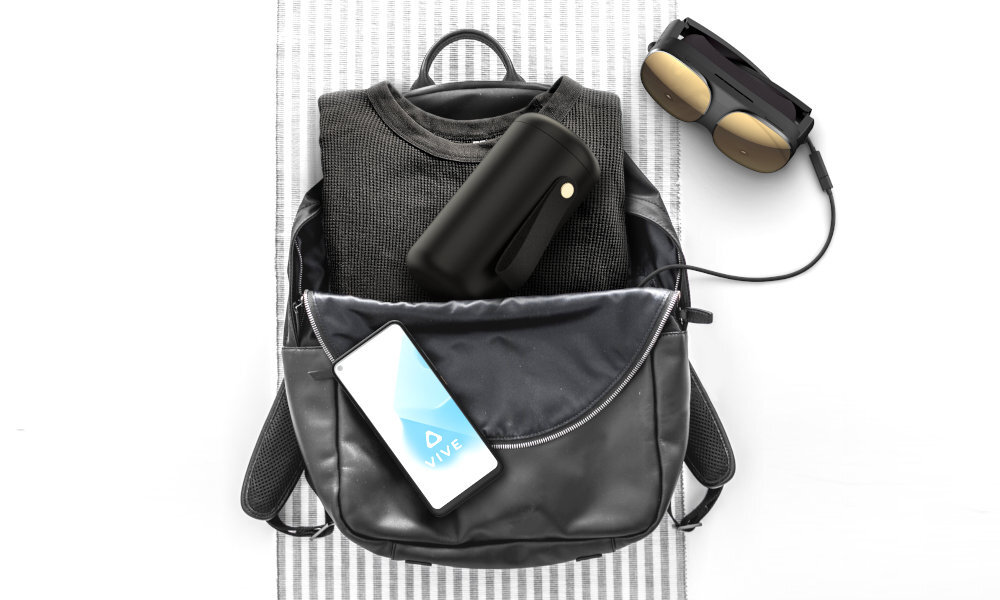 Na obrazie widoczny jest plecak z otwartą kieszenią, w której znajduje się czarny futerał i smartfon z logo \'VIVE\' na ekranie. Obok plecaka leżą gogle VR z podłączonym kablem.
