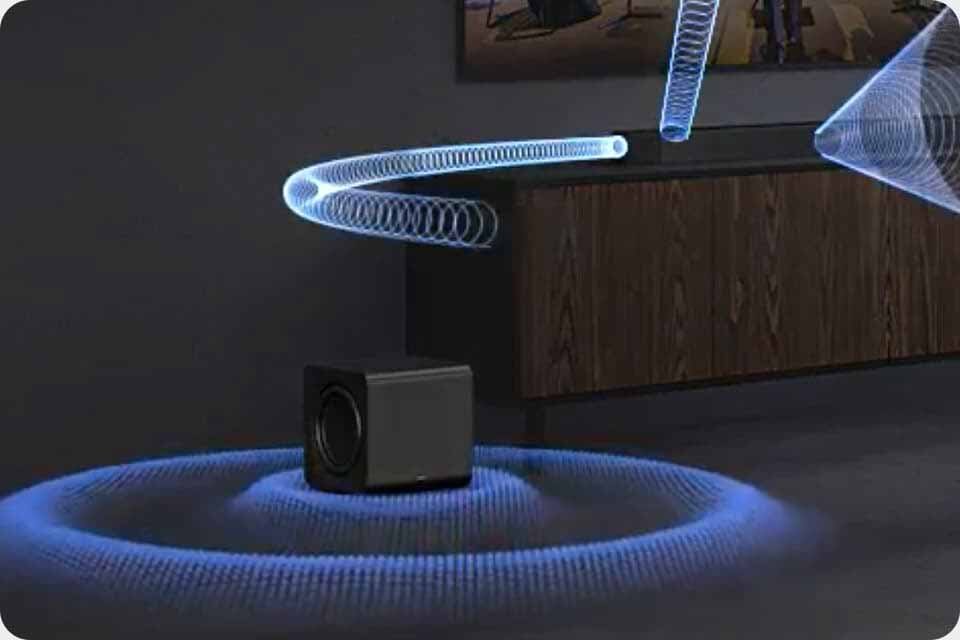 Na obrazie widać czarny subwoofer umieszczony na podłodze, z którego wydobywają się niebieskie fale dźwiękowe. W tle znajduje się drewniana szafka oraz fragment obrazu na ścianie.
