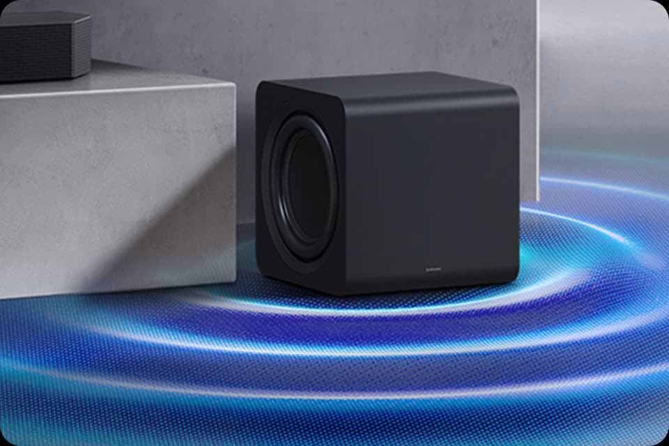 Czarny subwoofer o prostokątnym kształcie stoi na powierzchni z niebieskimi falami dźwiękowymi. Na subwooferze widoczne jest logo \'SAMSUNG\'.