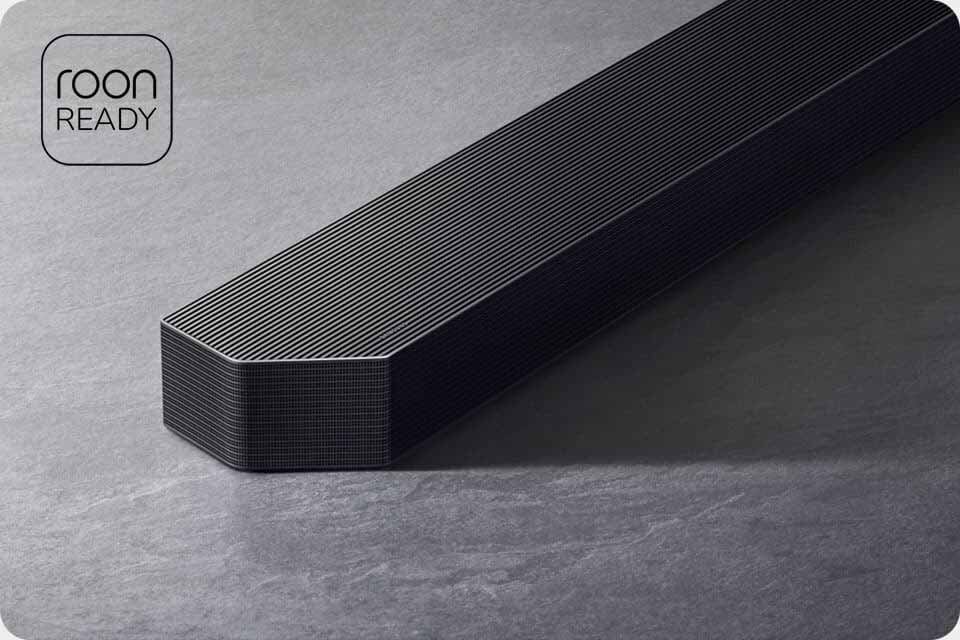 Na obrazie widoczny jest czarny soundbar o prostokątnym kształcie z zaokrąglonymi krawędziami, umieszczony na szarej powierzchni. W lewym górnym rogu znajduje się napis \'roon READY\'.