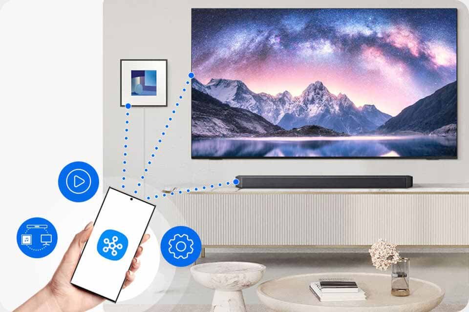 Na obrazie widać telewizor z krajobrazem górskim, poniżej którego znajduje się soundbar. W ręce trzymany jest smartfon z ikoną aplikacji, a wokół niego widoczne są symbole funkcji, takie jak odtwarzanie i ustawienia.