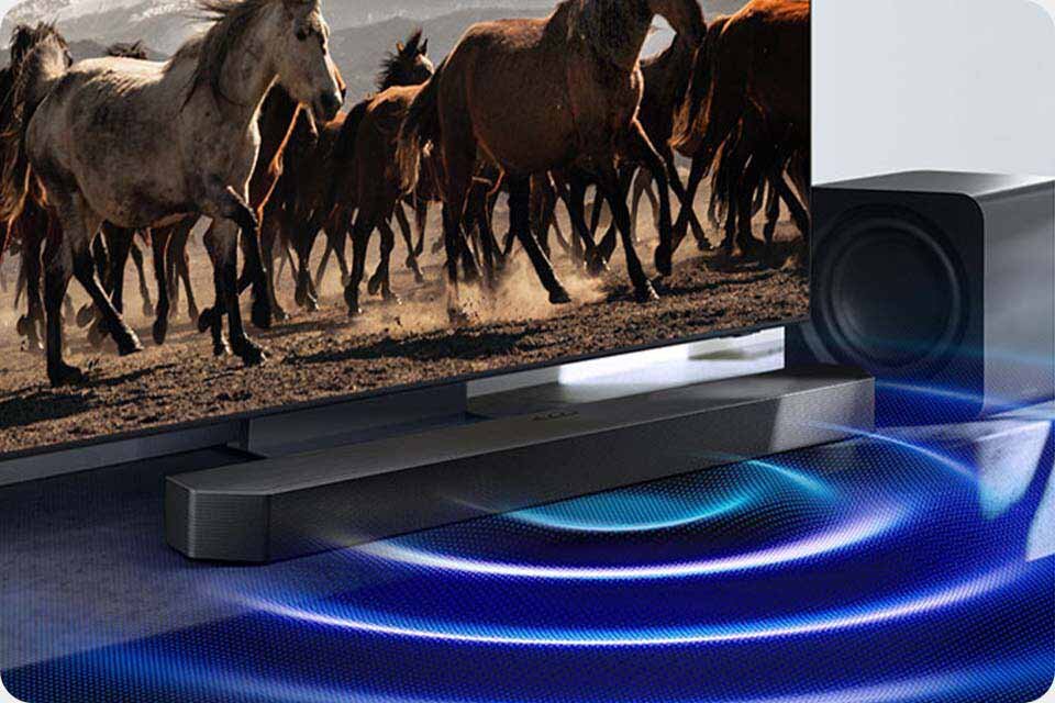 Na obrazie widoczny jest telewizor z wyświetlanym obrazem galopujących koni oraz czarny soundbar i subwoofer. Wokół soundbara widoczne są niebieskie fale dźwiękowe.