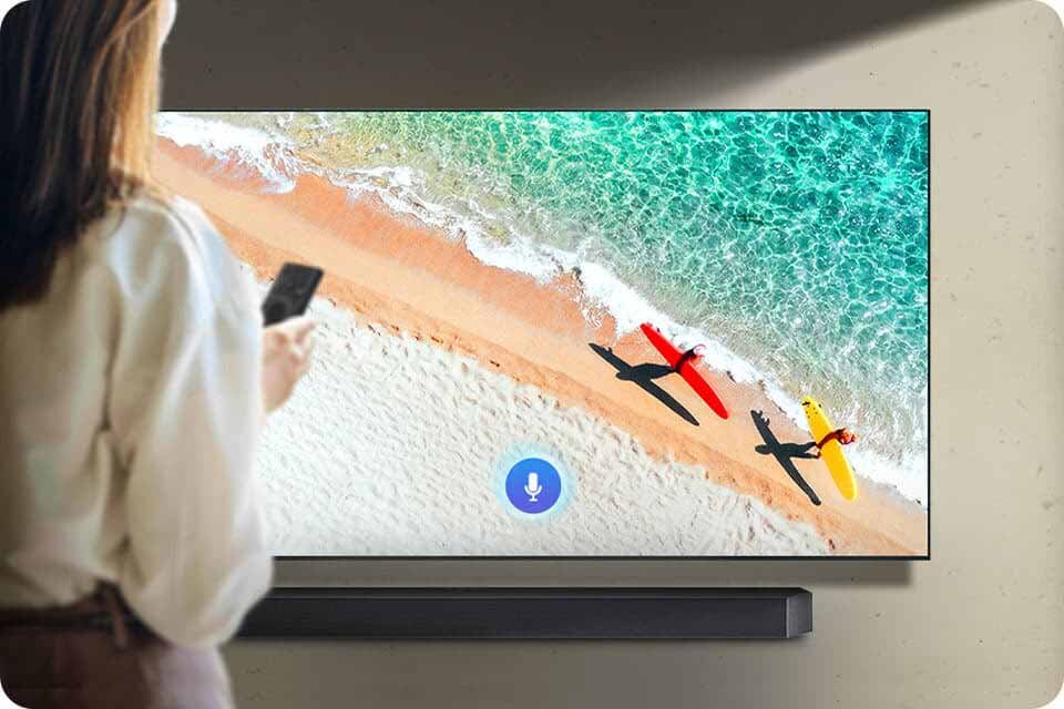 Osoba trzymająca telefon stoi przed dużym ekranem wyświetlającym obraz plaży z dwoma kolorowymi deskami surfingowymi. Pod ekranem znajduje się czarny soundbar.