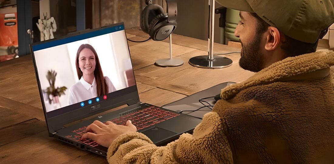 Osoba siedzi przy drewnianym stole, używając laptopa z czerwonym podświetleniem klawiatury, na ekranie widoczna jest wideorozmowa z kobietą. W tle znajdują się słuchawki na stojaku i inne przedmioty biurowe.
