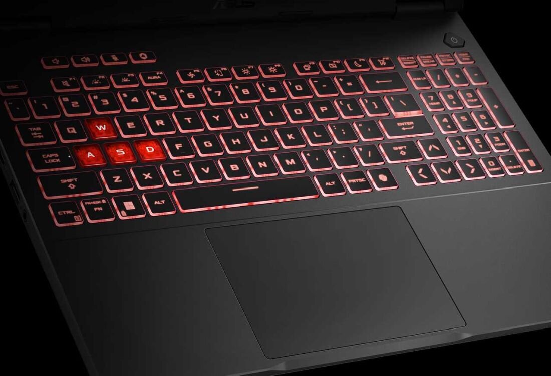 Klawiatura laptopa z podświetleniem w kolorze czerwonym, z wyróżnionymi klawiszami W, A, S, D. Widoczny jest duży touchpad poniżej klawiatury.
