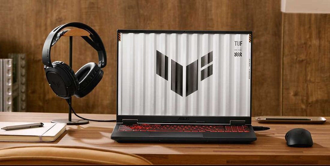 Na drewnianym biurku stoi otwarty laptop z widocznym logo TUF na ekranie. Obok laptopa znajdują się słuchawki na stojaku oraz mysz komputerowa.