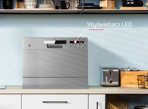 Na blacie kuchennym stoi srebrna zmywarka z widocznym wyświetlaczem LED. Obok znajduje się toster i koszyk z owocami, a nad zmywarką półka z naczyniami.