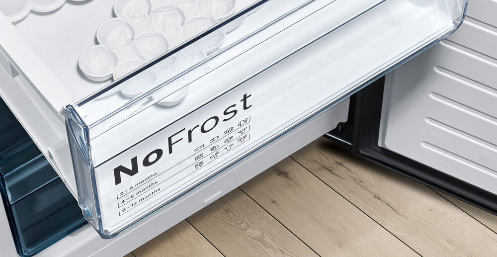 Otwarta szuflada lodówki z napisem \'NoFrost\' oraz ikonami i tekstem wskazującym na okresy przechowywania: 6, 12, 18 miesięcy. W tle widoczna jest drewniana podłoga.
