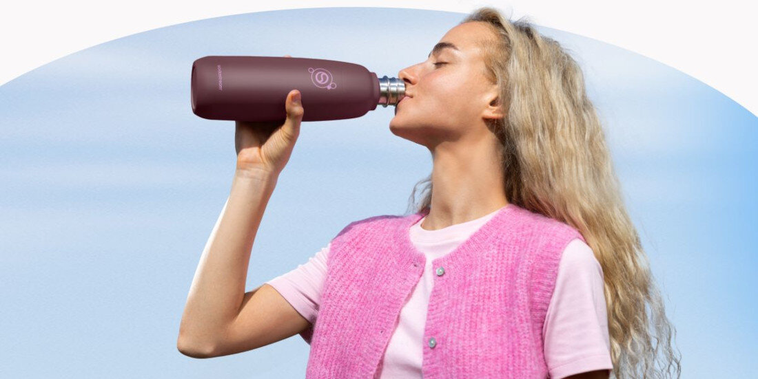 Osoba pije z bordowej butelki termicznej z logo SODASTREAM na tle nieba. Osoba ma na sobie różowy sweter i koszulkę.
