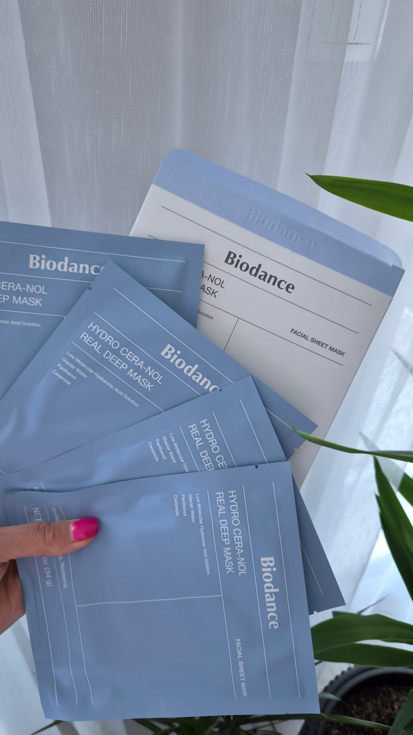 Na obrazie widoczne są cztery niebieskie opakowania z napisem \'Biodance Hydro Cera-nol Real Deep Mask\'. W tle znajduje się roślina i zasłona.