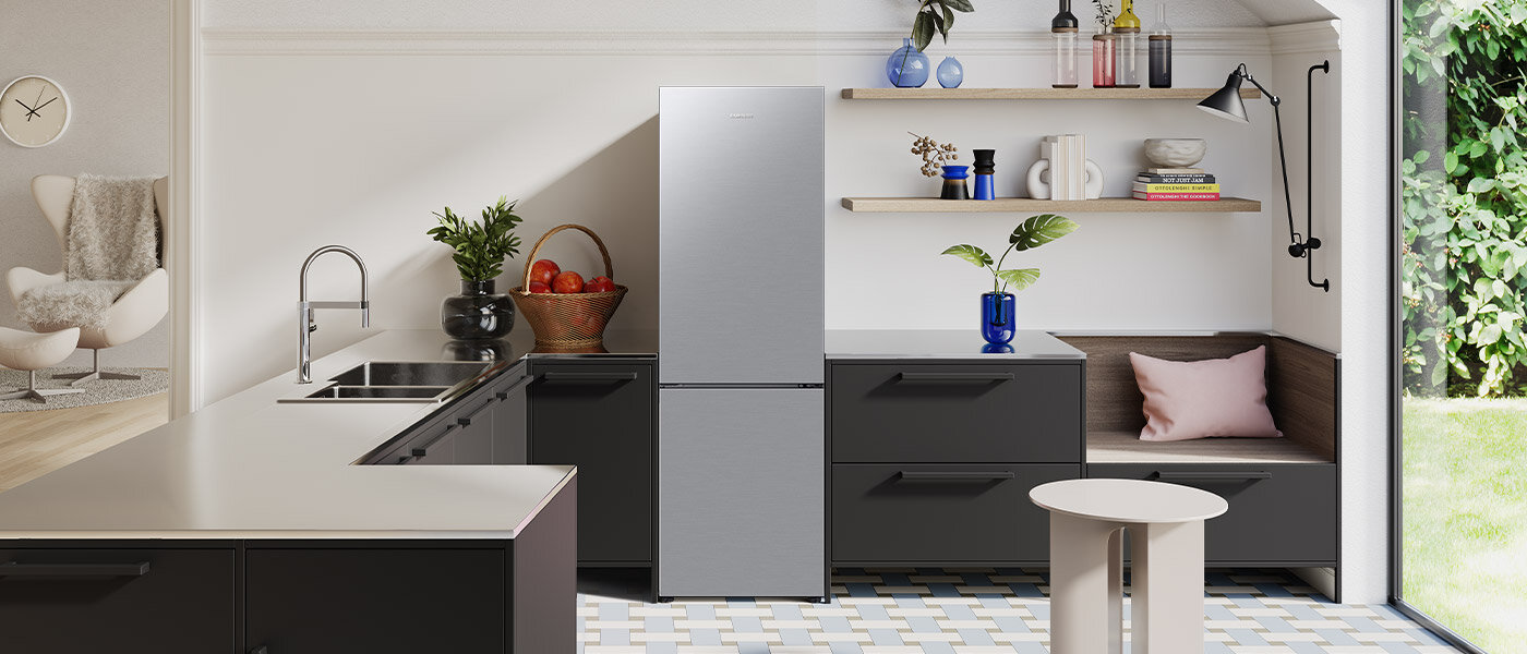 Elegancki widny salon z aneksem kuchennym widoczny na zdjęciu pokazuje dopasowanie estetyki lodówki Samsung do wnętrza współczesnego mieszkania Kuchnia z lodówką w metalicznym grafitowym kolorze, obok której znajduje się zlew i blat z owocami. Na półkach są dekoracje, a w rogu znajduje się siedzisko z poduszką.