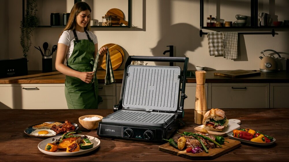 Na stole w kuchni znajduje się otwarty grill elektryczny z widocznymi potrawami wokół, takimi jak jajka sadzone, warzywa i kanapka. W tle stoi osoba ubrana w zielony fartuch, trzymająca ściereczkę.