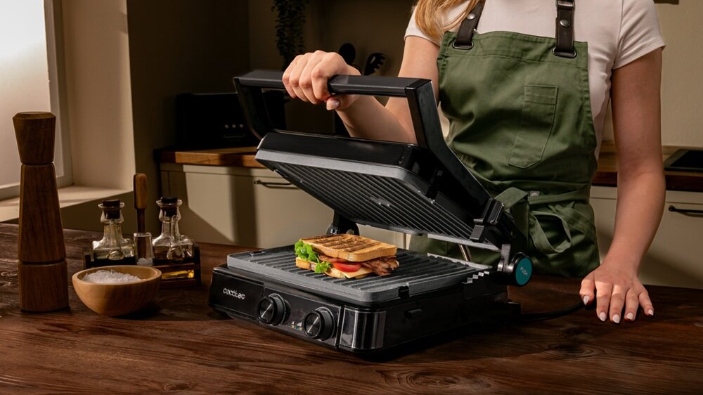 Osoba w zielonym fartuchu otwiera grill elektryczny, na którym znajduje się kanapka z warzywami. Na grillu widoczne jest logo \'CECOTEC\'.
