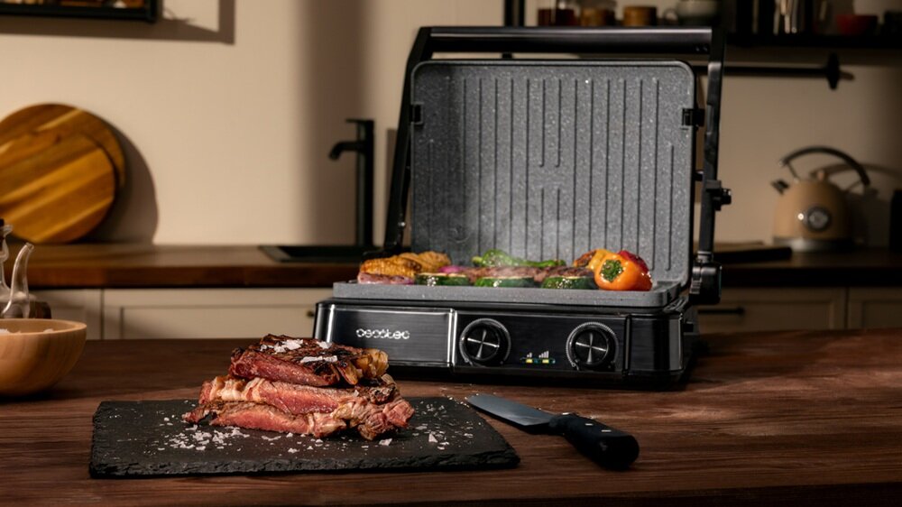 Na drewnianym blacie stoi otwarty grill elektryczny z jedzeniem na ruszcie. Przed grillem leży deska z kawałkami mięsa i nożem, a na grillu widoczne są warzywa i mięso.