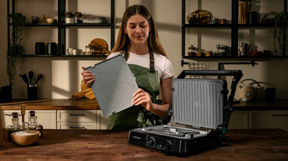 Osoba trzyma wyjmowaną płytę grilla elektrycznego CECOTEC Rock’nGrill 2200 Dual w kuchni. Na blacie widoczny jest otwarty grill z widocznymi elementami grzewczymi i logo CECOTEC.