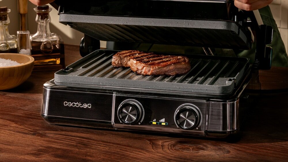Grill elektryczny z otwartą pokrywą, na którym znajduje się grillowany kawałek mięsa. Na przednim panelu widoczne są dwa pokrętła oraz logo \'cecotec\'.