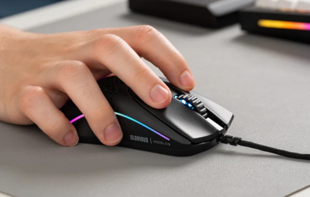 Osoba trzyma czarną mysz komputerową z podświetleniem RGB i napisem \'GLORIOUS MODEL O 2\'. Mysz jest podłączona kablem i znajduje się na szarej powierzchni.