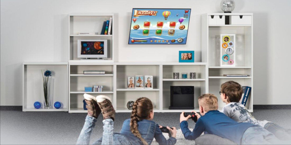 Trzech chłopców leży na podłodze, grając na konsoli przed telewizorem zamontowanym na ścianie. W tle znajdują się białe regały z książkami, zdjęciami i dekoracjami.