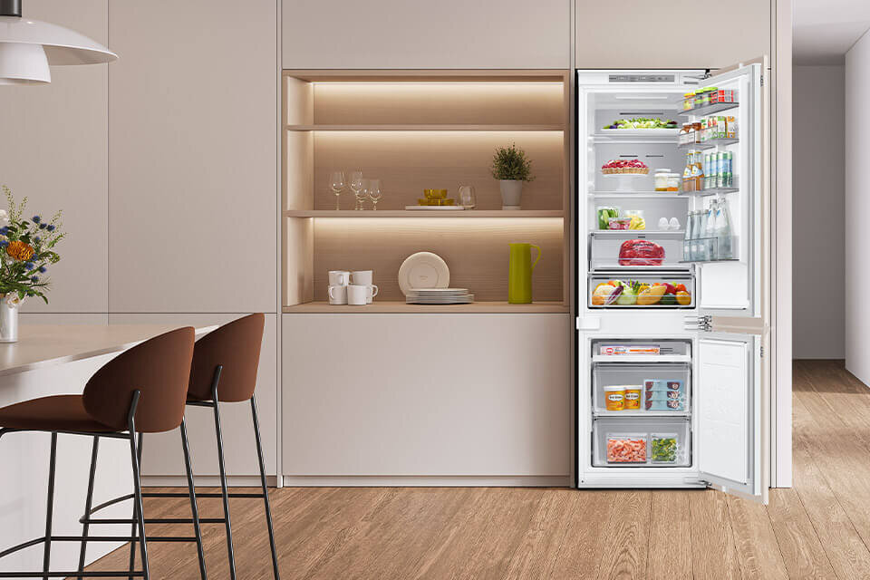 lodówka do zabudowy BRB80F26DEF0EO z oferty Media Expert Kuchnia z otwartą lodówką w zabudowie, wypełnioną różnorodnymi produktami spożywczymi. Obok lodówki znajdują się półki z dekoracjami i naczyniami.
