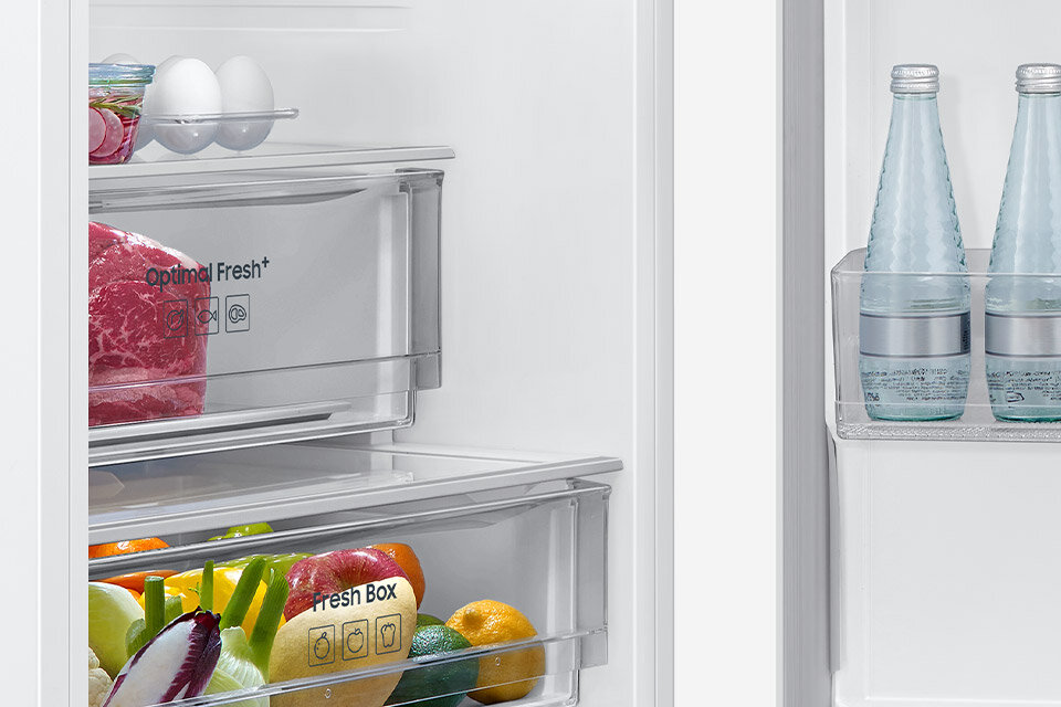 Wnętrze lodówki z widocznymi szufladami oznaczonymi jako \'Optimal Fresh+\' i \'Fresh Box\', wypełnionymi różnymi produktami spożywczymi. Na drzwiach lodówki znajdują się dwie butelki wody.