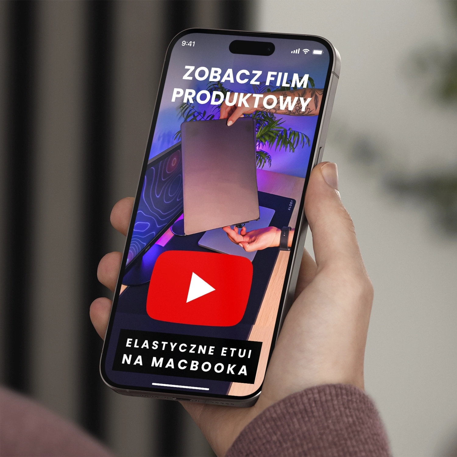 Osoba trzyma smartfon, na ekranie którego widoczny jest film z napisem \'ZOBACZ FILM PRODUKTOWY\'. Poniżej znajduje się duży przycisk z ikoną YouTube oraz napis \'ELASTYCZNE ETUI NA MACBOOKA\'.