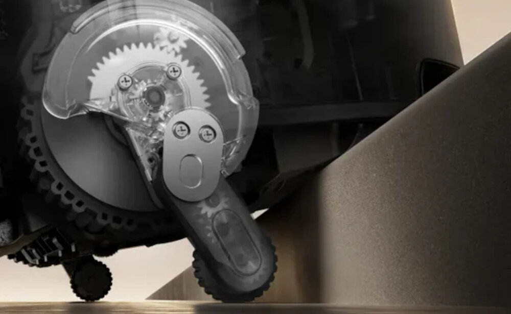 Zbliżenie na mechanizm zębatkowy robota sprzątającego, z widocznymi przezroczystymi elementami obudowy i kołami. Tło jest rozmyte, a robot znajduje się na krawędzi powierzchni.