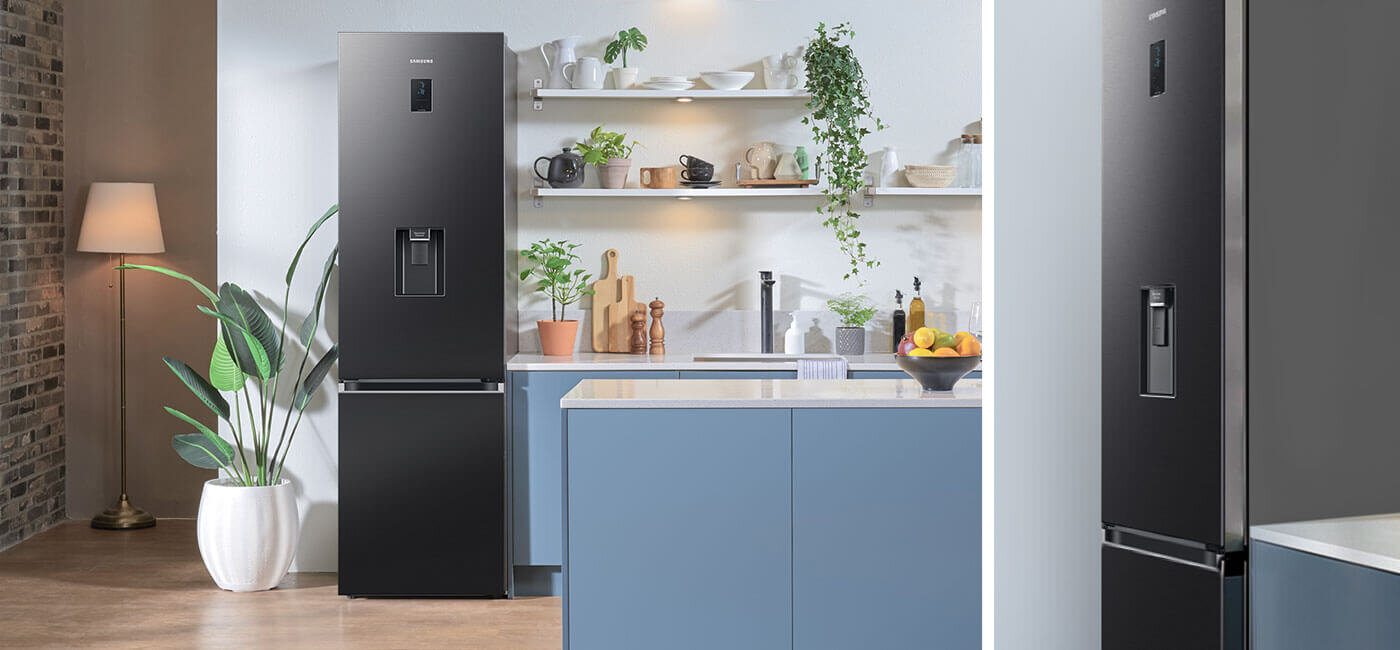 Design Samsung dopasowany do nowoczesnej kuchni Grafitowa lodówka z podajnikiem wody stoi w nowoczesnej kuchni z niebieskimi szafkami i roślinami na półkach. Na lodówce widoczne jest logo \'SAMSUNG\'.