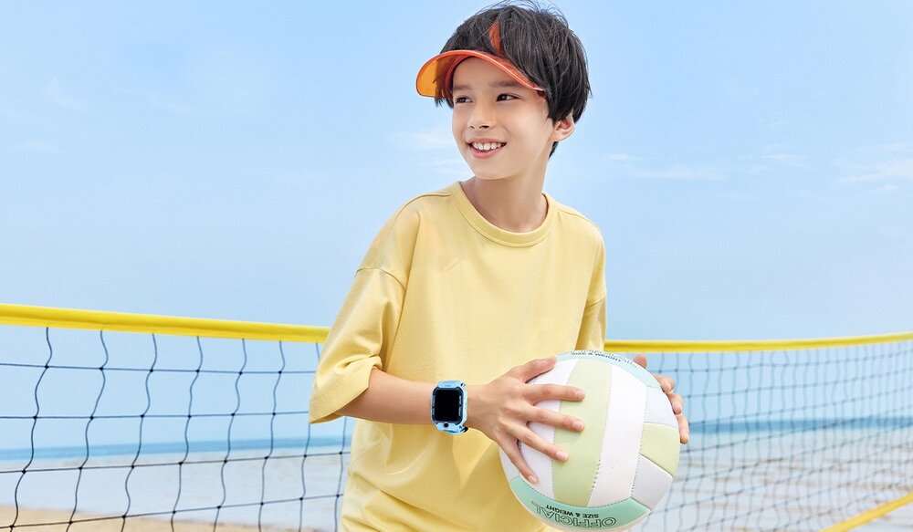 Dziecko w żółtej koszulce trzyma piłkę do siatkówki, na ręce ma niebieski smartwatch. W tle widać siatkę do gry i plażę.