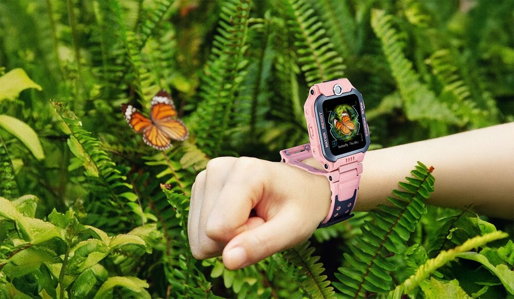 Na zdjęciu widoczny jest smartwatch na ręce, z wyświetlaczem pokazującym motyla. W tle znajdują się zielone liście paproci oraz motyl.