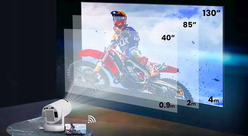 Obraz przedstawia projektor wyświetlający obraz motocyklisty na ekranie, z różnymi rozmiarami projekcji oznaczonymi jako 40”, 85”, 130”. Na podłodze obok projektora znajduje się smartfon z ikoną Wi-Fi.