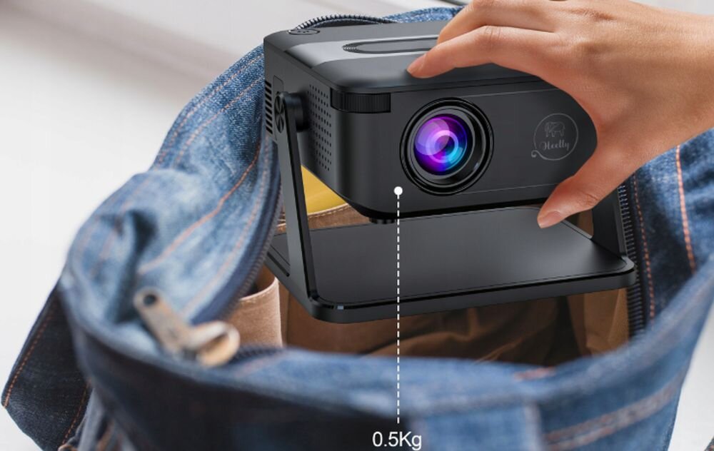 Osoba wkłada czarny projektor z widocznym logo \'Heelly\' do torby z dżinsu. Obok projektora znajduje się napis \'0.5Kg\'.