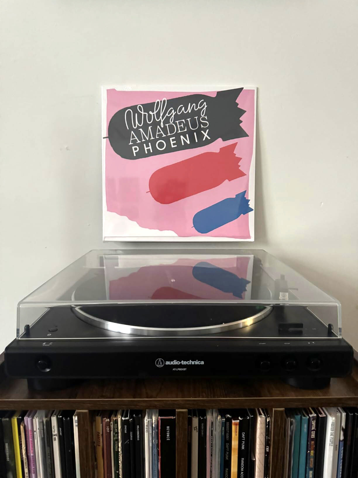 Na zdjęciu znajduje się gramofon z przezroczystą pokrywą, ustawiony na półce z płytami winylowymi. W tle widać okładkę albumu z napisem \'Wolfgang Amadeus Phoenix\'.