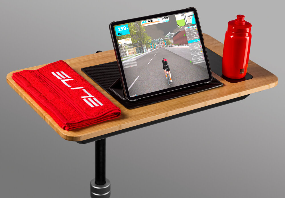 Na drewnianym blacie znajduje się tablet z wyświetlonym symulatorem jazdy na rowerze, obok czerwony ręcznik z napisem \'ELITE\' i czerwona butelka na wodę.