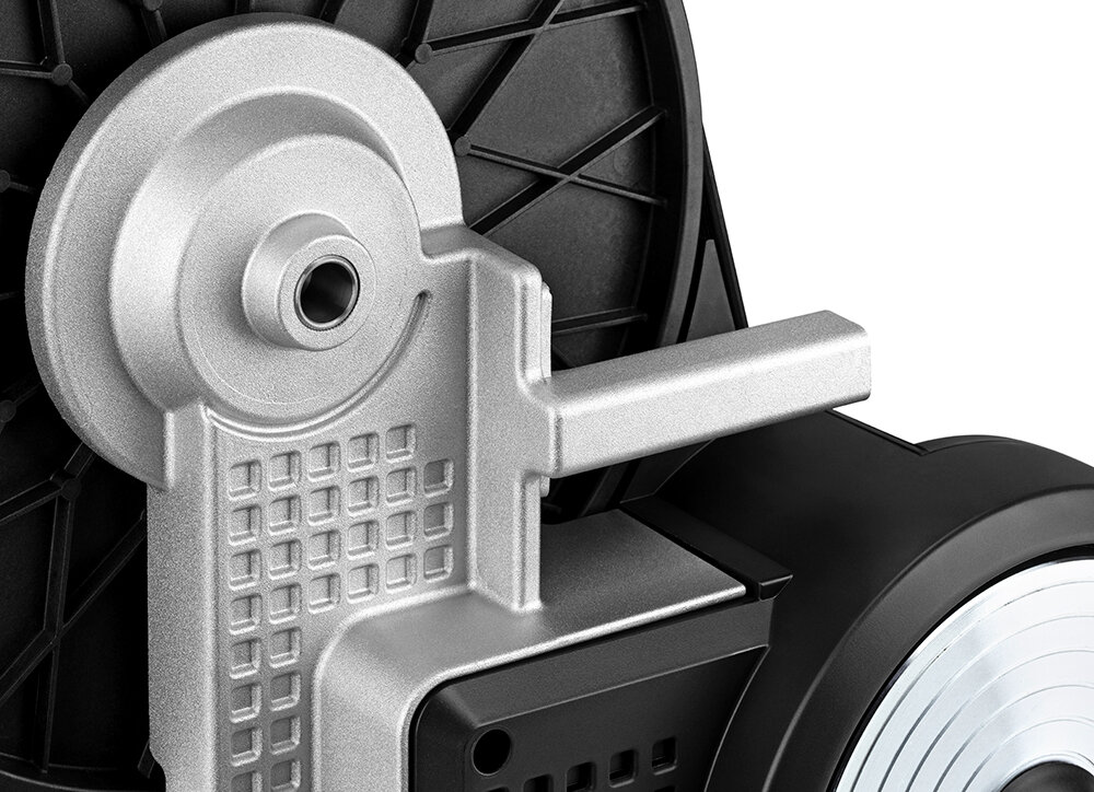Zbliżenie na metalowy element trenażera rowerowego z widocznymi detalami konstrukcyjnymi i otworem na śrubę. W tle widać czarną, plastikową część z wzorem kratki.