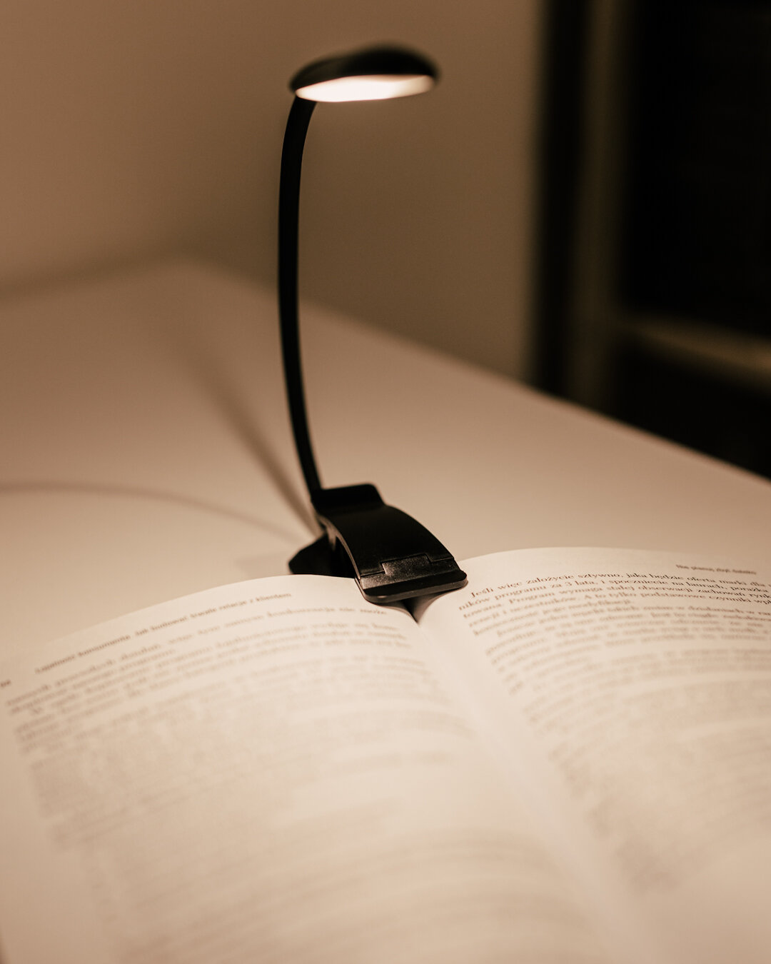 Lampka do czytania z klipsem oświetla otwartą książkę na biurku. Lampka ma elastyczne ramię i emituje ciepłe światło.