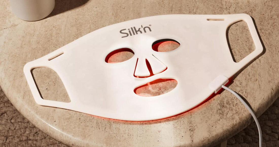 Na zdjęciu znajduje się biała maska LED do twarzy z wycięciami na oczy, nos i usta, leżąca na jasnym blacie. Na masce widoczne jest logo \'Silk\'n\', a z boku podłączony jest biały kabel.