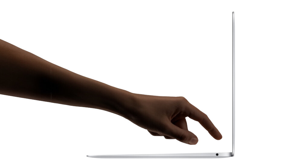 Na obrazie widać rękę dotykającą cienkiego laptopa, który jest otwarty pod kątem. Laptop ma srebrną obudowę i widoczny jest port na krawędzi.