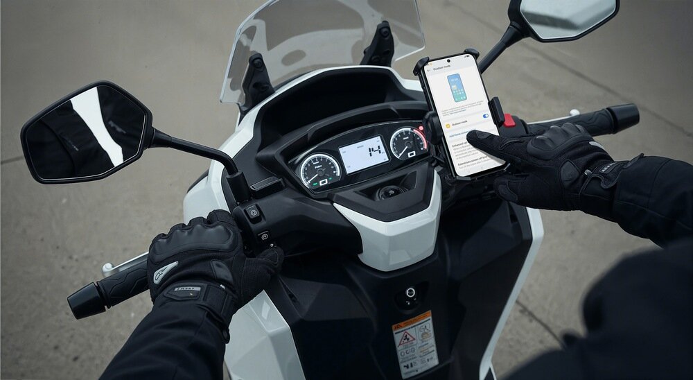 Osoba w rękawicach motocyklowych trzyma smartfon zamocowany na kierownicy skutera, na ekranie widoczny jest tekst i obrazek. Zegary skutera pokazują prędkość 14 km/h.
