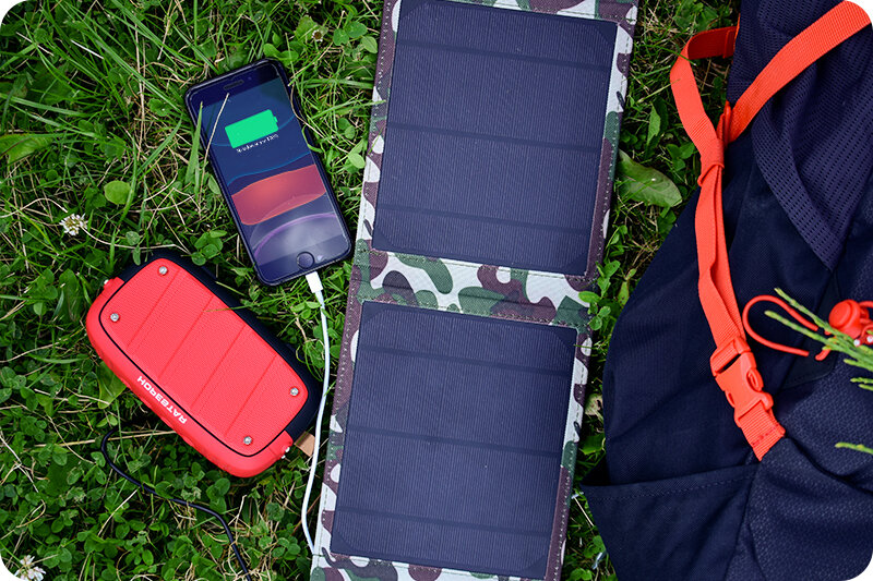 Na trawie leży turystyczna ładowarka solarna z trzema panelami, obok której znajduje się telefon z zieloną ikoną ładowania na ekranie oraz czerwony powerbank z napisem \'RATGEOH\'.