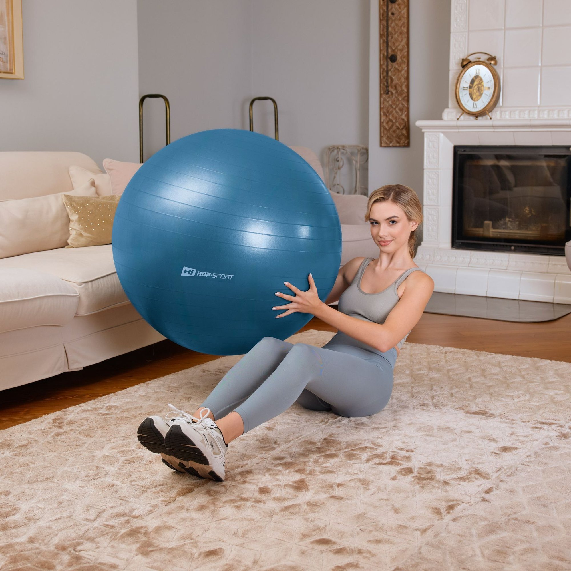 Osoba siedzi na dywanie w salonie, trzymając niebieską piłkę fitness z logo \'HOP-SPORT\'. W tle widać sofę, kominek i zegar na półce.
