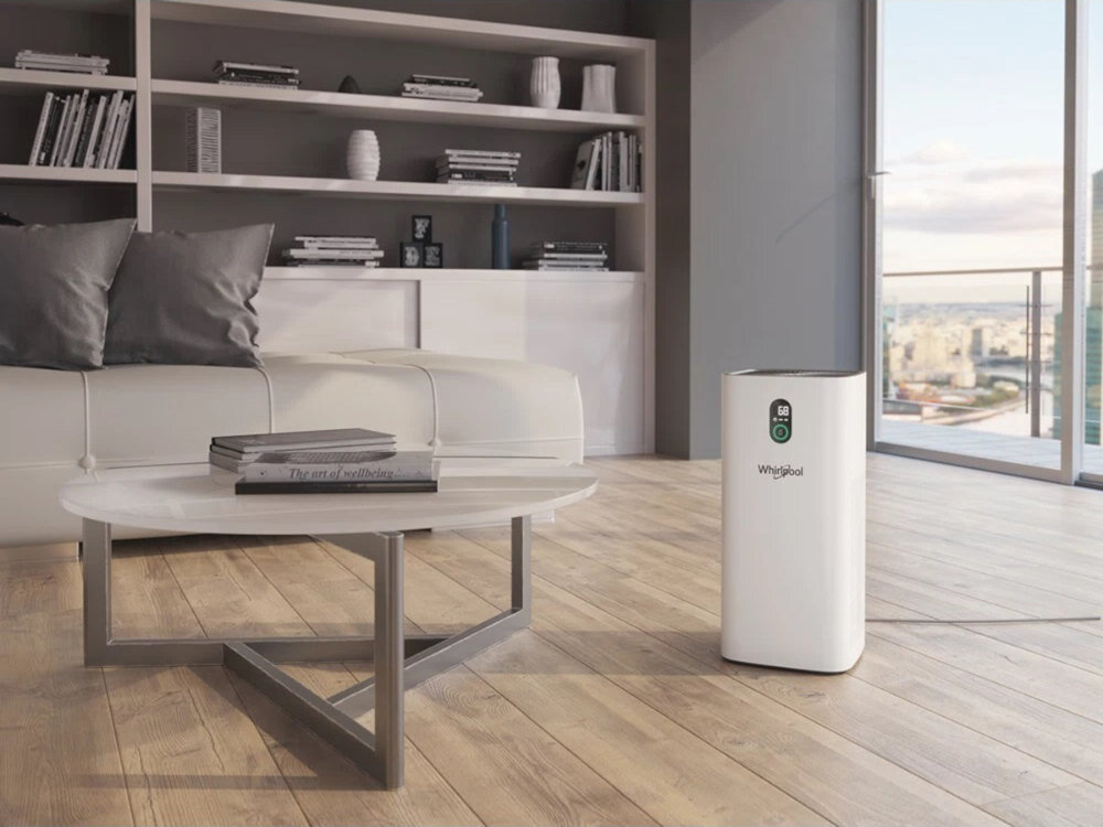 W nowoczesnym salonie z drewnianą podłogą stoi biały oczyszczacz powietrza z logo Whirlpool. W tle znajduje się biała sofa, stolik kawowy i regał z książkami oraz dekoracjami.