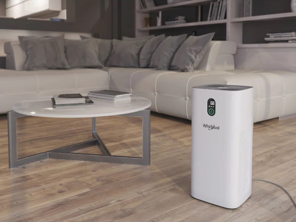 Na drewnianej podłodze stoi biały oczyszczacz powietrza z logo \'Whirlpool\', obok znajduje się biały stolik z książkami, a w tle widoczna jest jasna sofa z poduszkami.