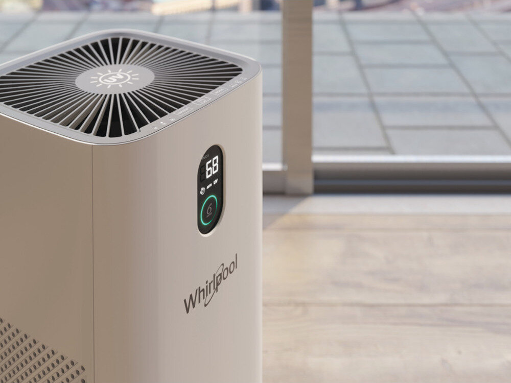 Na obrazie widoczny jest oczyszczacz powietrza z wyświetlaczem pokazującym liczbę 68 oraz logo Whirlpool. Urządzenie stoi na podłodze przy oknie, a na górze ma kratkę wentylacyjną z symbolem słońca.