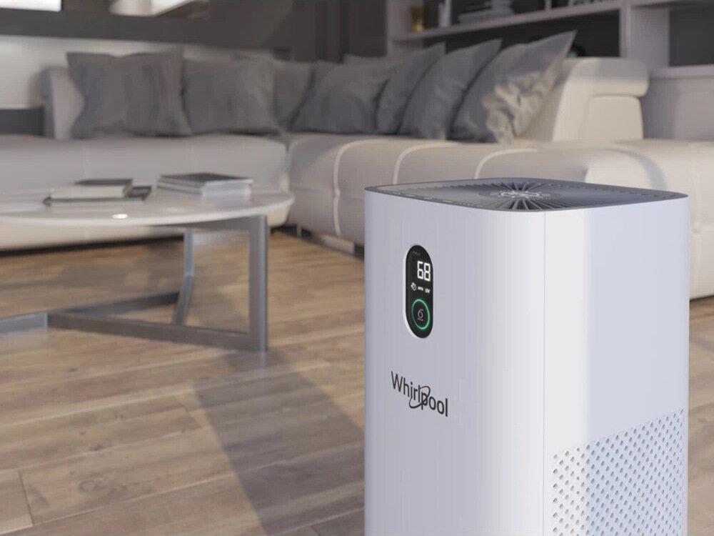 Na pierwszym planie znajduje się biały oczyszczacz powietrza z wyświetlaczem pokazującym liczbę 68 i logo Whirlpool. W tle widoczna jest jasna sofa i stolik w nowoczesnym wnętrzu.